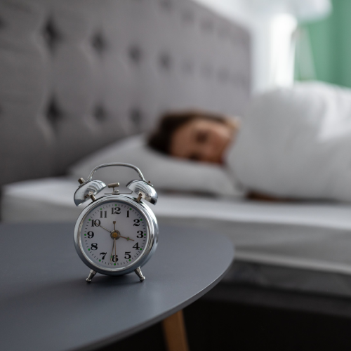 Silberner Wecker auf einem Nachttisch im Vordergrund, zeigt 6:30 Uhr. Im Hintergrund liegt eine Person unter einer weißen Bettdecke im Bett und schläft.