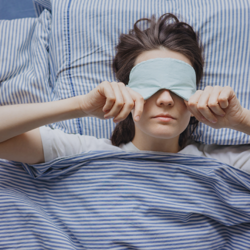 Frau liegt im Bett mit blau-weiß gestreifter Bettwäsche und zieht sich eine hellblaue Schlafmaske über die Augen, bereit zum Einschlafen.