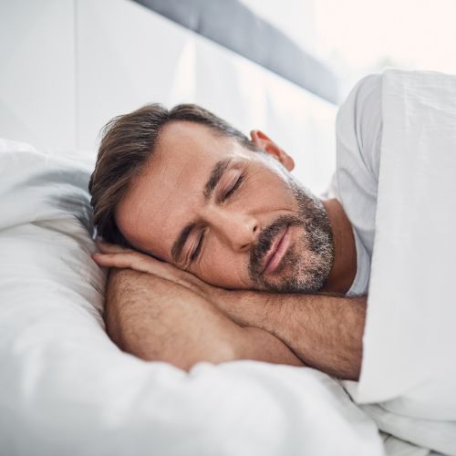 Ein Mann mit Bart liegt seitlich im Bett und schläft entspannt, den Kopf auf seine Hände gebettet und zugedeckt mit einer weißen Decke.