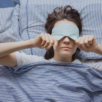 Frau liegt im Bett mit blau-weiß gestreifter Bettwäsche und zieht sich eine Schlafmaske über die Augen.